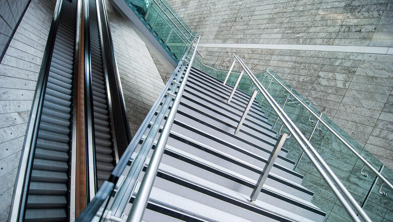 Foto: iStock.de/Ilbusca Das Foto zeigt zwei Rolltreppen sowie eine Treppe, die über die beiden Rolltreppen führt. Die Rolltreppen laufen gerade aus. Die moderne aus Metall bestehende Treppe überquert die Rolltreppen, jedoch versetzt im Zickzack.