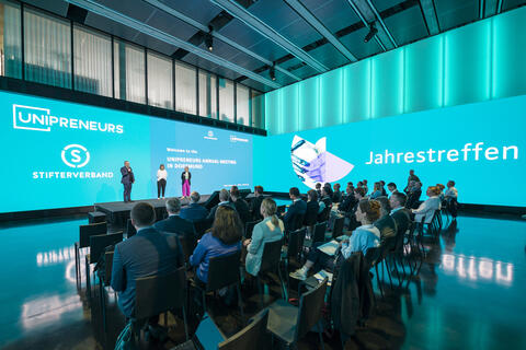 Begrüßung auf der Jahrestagung der Initiative Unipreneurs. Blick auf die Bühne. Im Vordergrund sitzt das Publikum